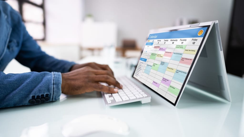 A person types on a wireless keyboard at a desk while viewing a digital calendar filled with colored events on therapy scheduling software displayed on a convertible laptop in tent mode.